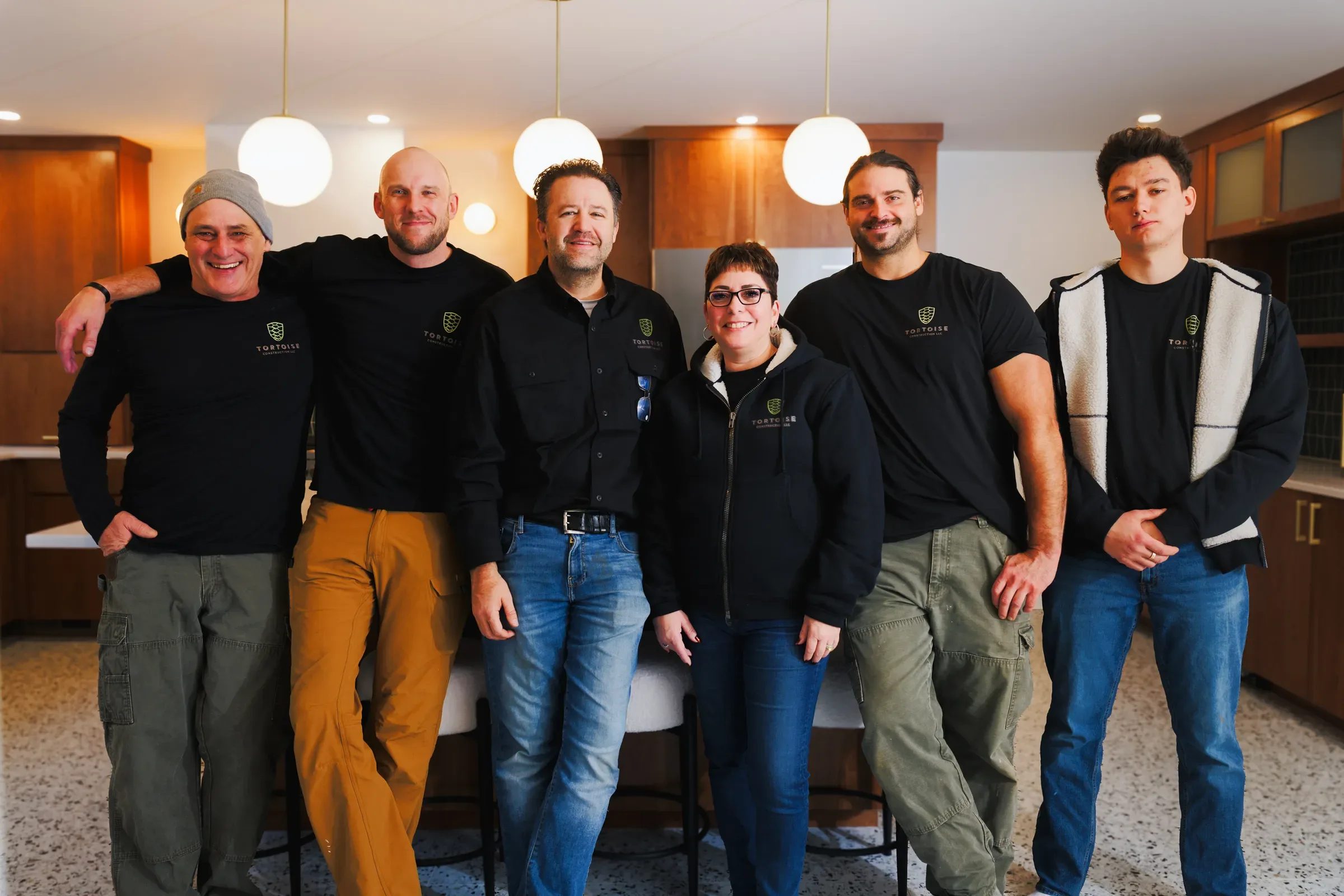 The Tortoise Construction crew of six in branded shirts, standing together in a remodeled kitchen with walnut cabinetry and globe pendant lights